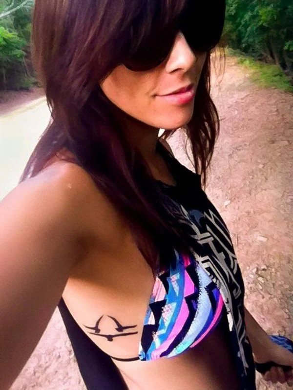 Mujer morena en el bosque, lleva gafas de sol y sonrie, tiene en sus costillas tatuaje con tres palomas formando signo de libra