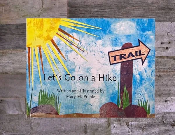 cover of children's book, Let's Go on a Hike, features handmade collage art illustration of hiking trail with sun, rocks, greenery, and trail marker
