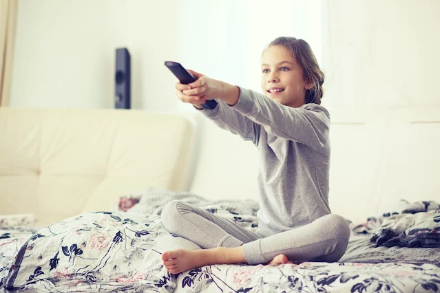 child sitting on bed with a tv remote watching tv in bedroom