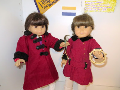 Two American Girl dolls in red coats holding musical instruments, left doll holding brass triangle and right doll holding tamborine