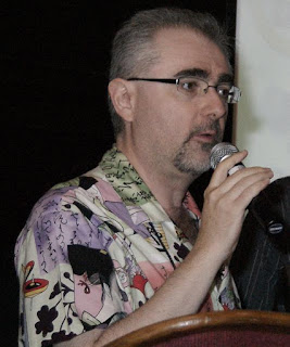 Robin Laws, an older gentleman with short grey hair, a thin goatee and moustache, and black glasses, wearing a colourful shirt, speaking into a microphone.