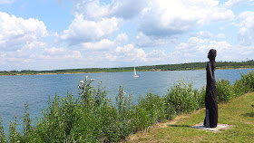 Das Foto zeigt den Zwenkauer See bei Leipzig bei Sonnenschein mit wenig Wolken. Ein weißes Segelboot ist auf dem See zu erkennen. Am Ufer wachsen Büsche und eine Holzskulptur in Form einer Frau ist zu erkennen.