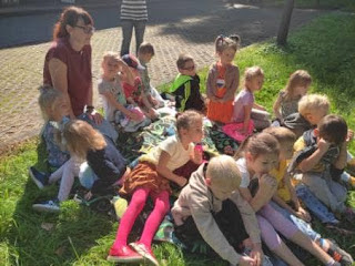 Pani przedszkolanka z grupą przedszkolaków siedzi na kocu na trawie.