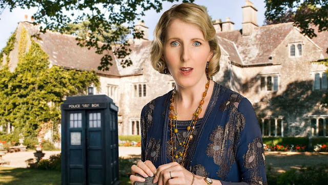 A white woman with short blonde hair wearing a blue 1920s style dress in front of a stately home covered in greenery. Behind her is the blue TARDIS.