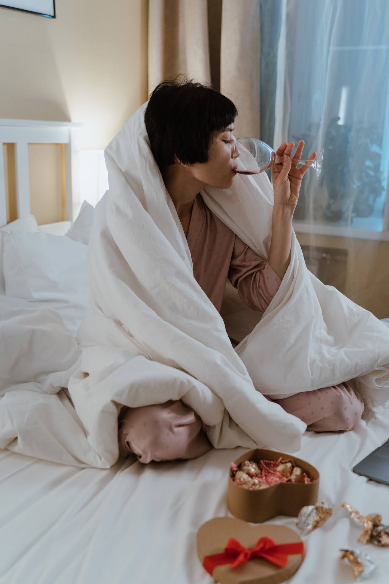 Woman drinking wine on a bed with chocolates nearby. Image by Pexels