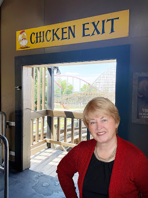 Janet, a light-skinned woman with light brown hair in a red shirt stands in a doorway with a rollercoaster in the background. Above the door-frame is a yellow sign that reads "Chicken Exit."