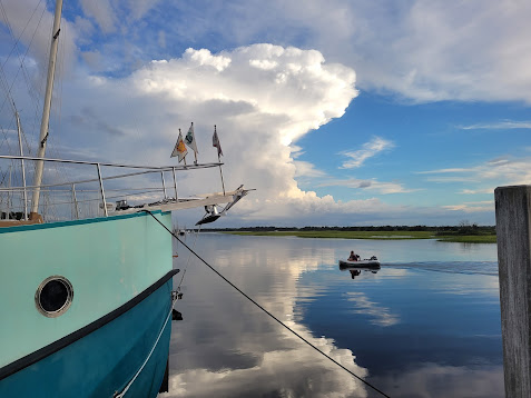 The bow of a teal trawler yacht is in the foreground.  A dinghy slices through the glassy water behind it, while a massive thundercloud rises into the sky, lit by the setting sun.