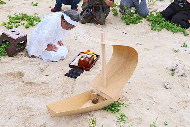 Priestess bowing and praying, photographers on her left, miniature sailboat