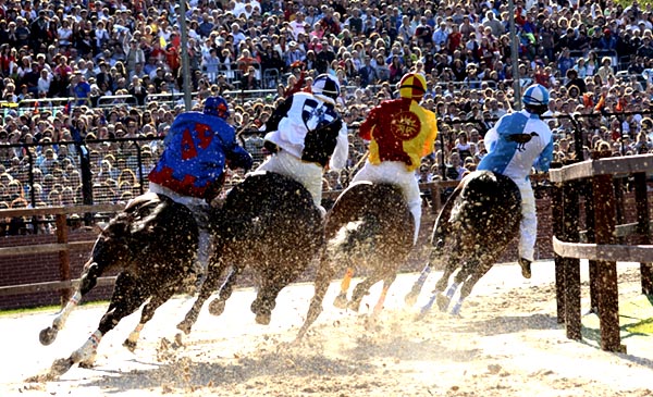 four horses rounding a curve along a wooden rail fence, all riders in jockeys' silks