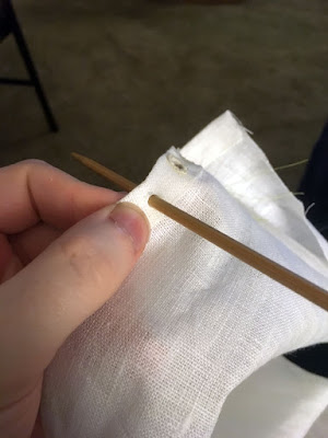 A close-up of a white finger and thumb holding a piece of white linen, with a wooden knitting needle inserted through the narrow double-folded hem.