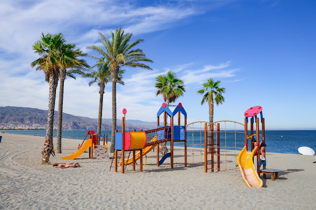 Parque infantil y palmeras sobre la arena de la playa con las azules aguas del mar al fondo