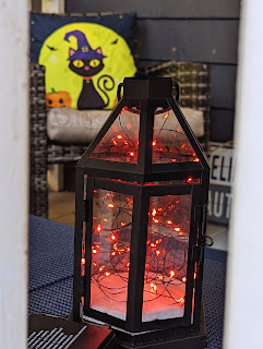 In the foreground, a lantern with red lights and behind it, a chair with a a pillow that has a black cat with witch's hat standing in front of the moon