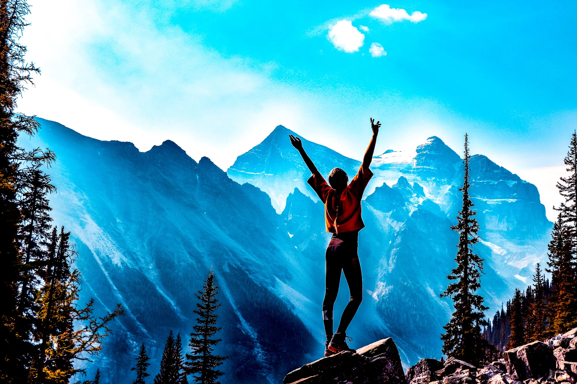 Someone stands on a mountain rock in a natural setting with arms raised, connecting with the natural world as part of their physical health journey.