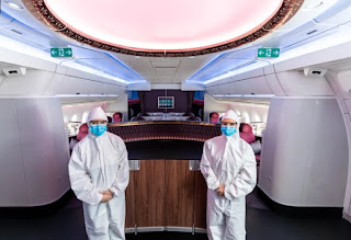 Foto no interior de uma aeronave da Qatar Airways. Em pé e de frente, lado a lado a uma distância aproximada de 2 metros, com os braços lateralizados e as mãos dadas sobre o abdome, dois comissários de voo, posicionados de costas para a divisória da primeira classe. Ambos usam macacão e touca em branco e máscara azul cobrindo nariz e boca. Ao fundo, atrás da divisória, a área preenchida por oito espaçosas poltronas. O lounge é amplo, os bins (compartimentos de bagagem acima das poltronas) possuem acabamento arredondado. A parte superior dos bins possui um design arrojado e futurista, contrasta com a iluminação do teto de luz branca embutida em uma moldura circular e clássica. Em ambos os lados, fixados acima dos bins, duas sinalizações em verde de saídas de emergência, uma de cada lado.  Com esta imagem desejamos um Feliz Dia do Comissário de Voo!