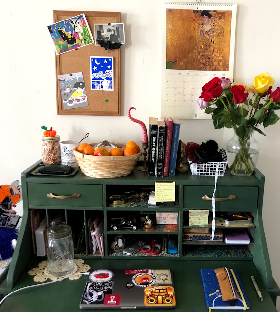 A dark green secretary desk with gold handles and spaces full of papers, cluttered with objects from college life including a laptop, jar of water, basket of oranges, books and a bouquet of red and yellow roses. above the desk is a calendar with art by Gustav Klimt, and a bulletin board with postcards from friends.
