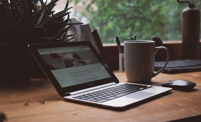 An open laptop, mouse and ceramic mug sit on a wooden desk