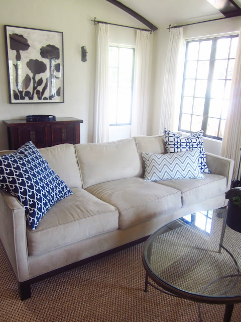 beige sofa with COCOCOZY accent pillows, oval glass table with metal legs on a woven rug. Behind it is a modern flower print and a small cabinet.