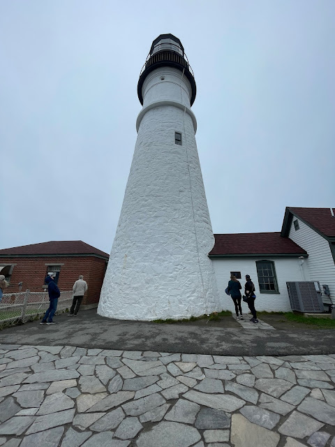 A close up of the lighthouse. It is white painted rock with a black roof. There are small gardens of flowers surrounding the lighthouse as well as a stone patio.