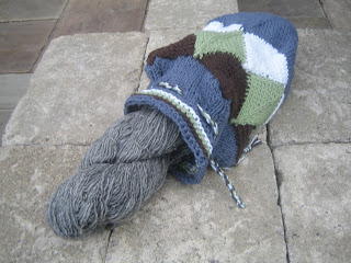 An image of a knitted bag, made up of mitered squares knitted in four colours.  It is laying flat, and open.  The top has eyelets and a drawstring cord. 