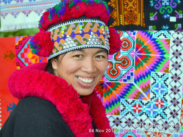 Matt Hahnewald; Facing the World; Asia; Northern Thailand; Lu-mien; Yao; travel destination; tourism; hilltribe; ethnic minority; ethnic; traditional Yao costume; traditional Yao headdress; Chiang Rai; Ban Paoo Naglae; woman; world cultures; red pompoms; smiling; eye contact; street portrait; closeup; Nikon DSLR D3100; 50 mm prime lens; three-quarter view; over-the-shoulder look