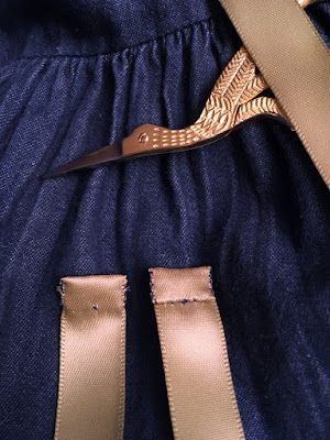 A closeup of small gold-handled stork scissors on a gathered denim background, with pale sage-green satin ribbon looped over them. Two ends of ribbon, folded over and stitched with blue thread, protrude from the bottom edge of the photo.