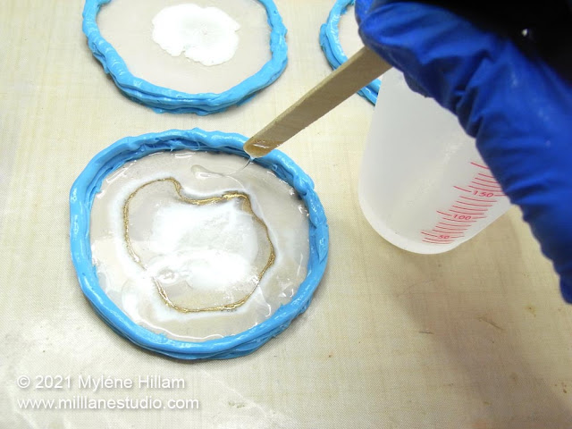 Gloved hand drizzling clear resin into a circular silicone mould over white resin to create an agate effect
