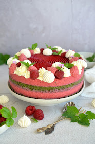 Himbeer-Dirndlmousse-Torte mit Brennnessel-Biskuit – zu sehen sind die Schichten aus grünem Brennnessel-Biskuit und rosarotem Dirndlmousse sowie die Dekoration aus Schlagobers, Himbeeren, Baiser, Blättern und Blüten an der Oberseite.