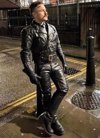 Leatherman with a mustache wearing full black leather gear in against a pole outside