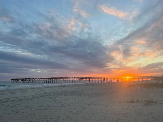 The beginning of the sunset taken from afar. The sun has just come down from the clouds that were blocking its descent. The sky has not yet turned its kaleidoscope of colors yet. The sky is filled with dark gray clouds, the beach and the water are calm.