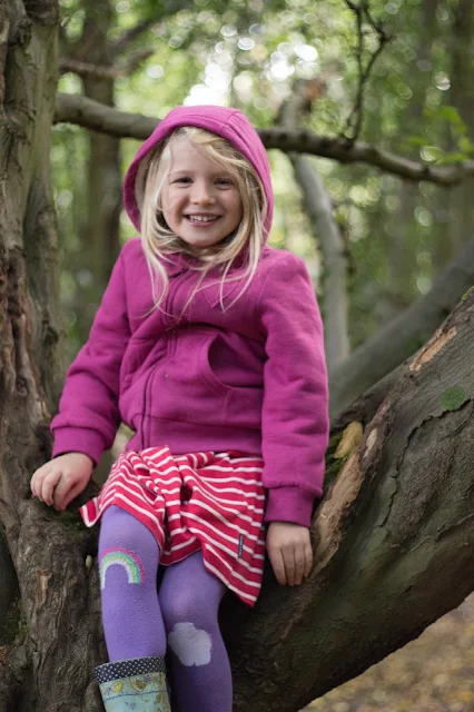 A 5 year old girl in a pinky hoody sits smiling in a tree