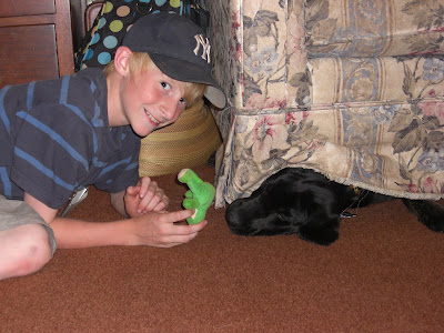 Picture of one farm camper smiling at the camera while holding Rudy's toy. Rudy's head is sticking out from under the couch trying to reach the toy