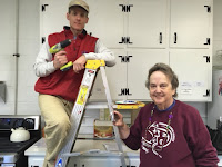 a man with a drill stands on a ladder while a woman smiles at the camera