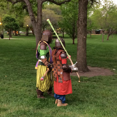 Two people in medieval clothing and armour, in a verdant park. They're facing slightly away from the camera, each holding a rattan sword. The nearer one is in red and blue, with a leather armour coat and a long red braid, and the further in green with a purple and gold surcoat belted on.