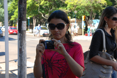 Tania está centralizada na foto, usando blusa vermelha, óculos escuros e segurando a câmera. Ao fundo aparece uma mulher que estava caminhando na calçada.