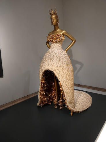 Photo of a dress on a mannequin. The mannequin is gold-colored. The dress is sleeveless and strapless, with a bodice of gold cloth flowers. The rest of the dress is floor-length and bell-shaped, very structured and made of gold-embroidered heavy cream fabric. It has a short train. In front, there's a cutout revealing the mannequin's legs and showing the inside of the skirt, which is lined with metallic gold fabric.