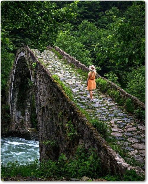 Ponte de pedra, por entre as árvores, sobre ela uma jovem caminhando solitária.