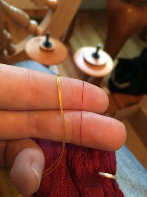 A close-up of doubled gold thread and a single strand of extremely fine deep garnet thread looped over the first two fingers of a left hand, in front of two spare bobbins on a spinning wheel's lazy kate. A tuxedo cat is blurrily visible under the table whose near leg is just in frame.