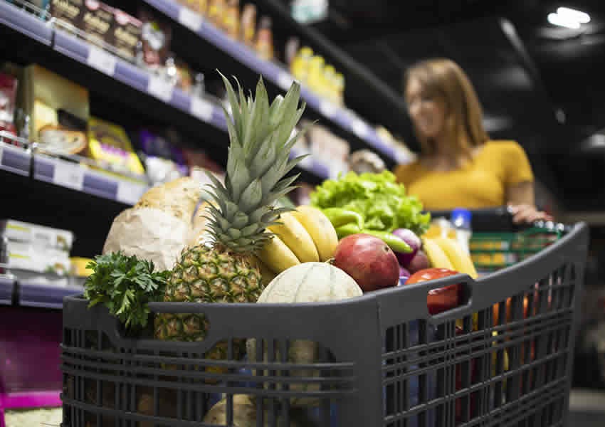 Imagem mostra uma mulher entre os corredores de um supermercado fazendo a compra da cesta básica perto de um carrinho de compras já lotado de alimentos.