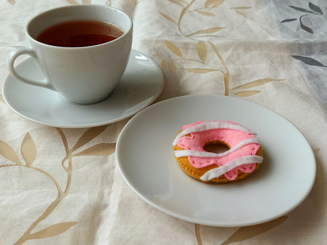 Filcowy donut leżący na talerzyku. Obok stoi filiżanka herbaty.