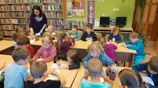 tło: regały z książkami. Panie bibliotekarka wśród przedszkolaków siedzących przy stolikach.