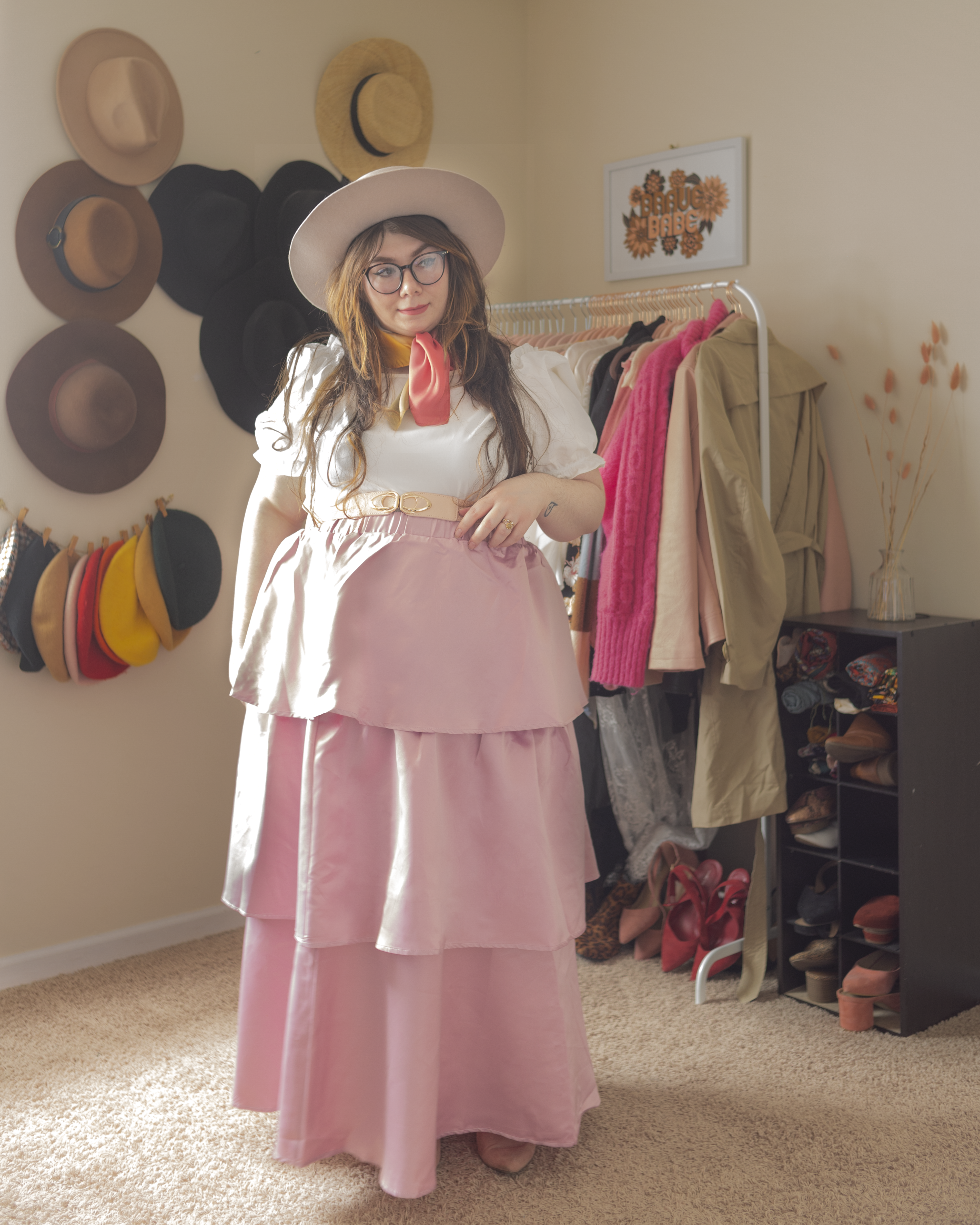 An outfit consisting of a beige and pink porkpie hat, pink and orange neck scarf tied around the neck, white puffy sleeve crop top paired with a pastel pink tiered maxi skirt, and pink mules.