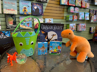 a green felt bucket with a dinosaur face sits next to a ball, a small toy dragon, a book about a dragon, and an orange plush dinosaur