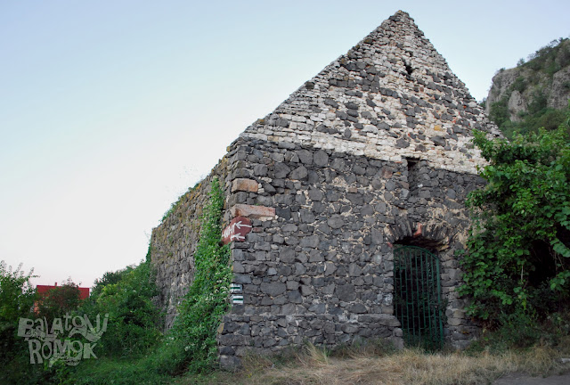 Bazaltkőből rakott templomfal, a nyugati ajtónyílással. A fal helyenként borostyánnal benőve. Jobbra a Csobánc bazaltormai.