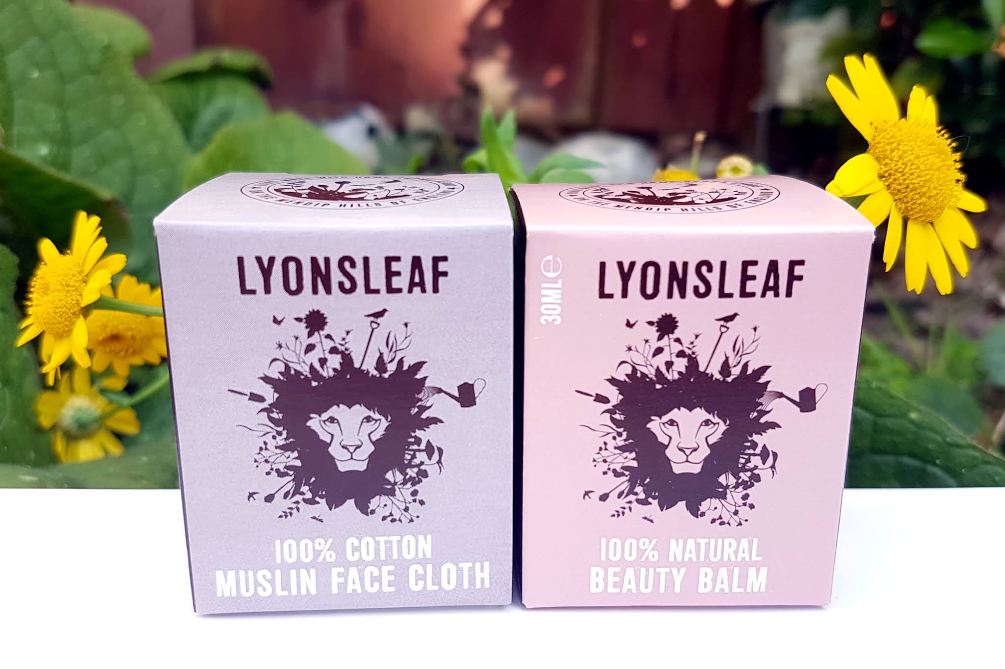 Lyonsleaf Beauty Balm and muslin cloth in purple and pink boxes sitting amongst yellow flowers