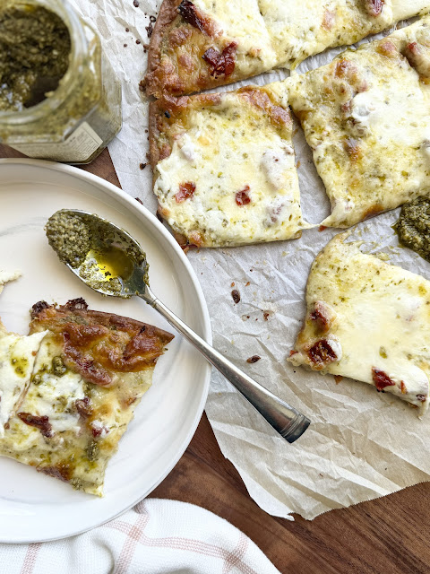A jar of basil pesto next to a slice of pizza on a white plate.