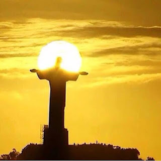 Foto em fundo dourado ao entardecer da gigantesca escultura do Cristo Redentor com os braços abertos abençoando a cidade do Rio de Janeiro, sob o intenso brilho do perfil da lua cheia que circunda a cabeça e parte dos braços do Cristo como se fosse uma enorme auréola. 