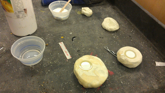 On a grey cart sits two round chunks of clay about the size of an adult's palm and a child's palm.  In each is a dome shaped dent with a diameter of about one inch.  There is a clear plastic starting to solidify from a syrupy liquid the middle of the clear plastic is turning a milky white.  Behind the clay is a white bottle and a couple of small plastic clear cups.