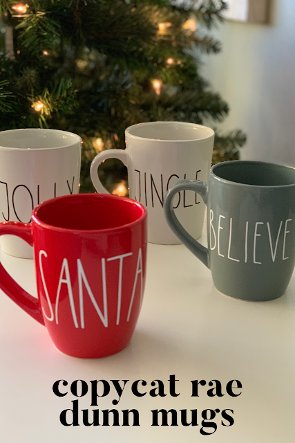 Four mugs, two white, one red and one gray that say Jingle, Believe, Jolly and Santa sitting on a white table by a lit Christmas tree.