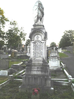 A tall pillar style gravestone, square in section and made of white stone.  It rises from a tierd base to a square column with writing insised in an arched panel.  Upwards the column tapers to a plinth which is surmounted by a draped vase.