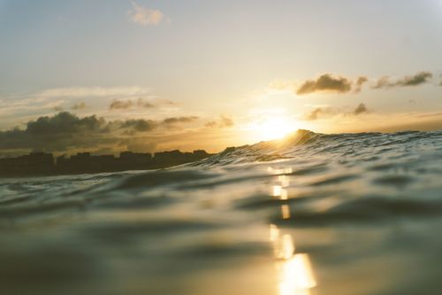 Fotografia de uma onda se formando, tirada de dentro do mar. O sol se põe no fundo da cena.